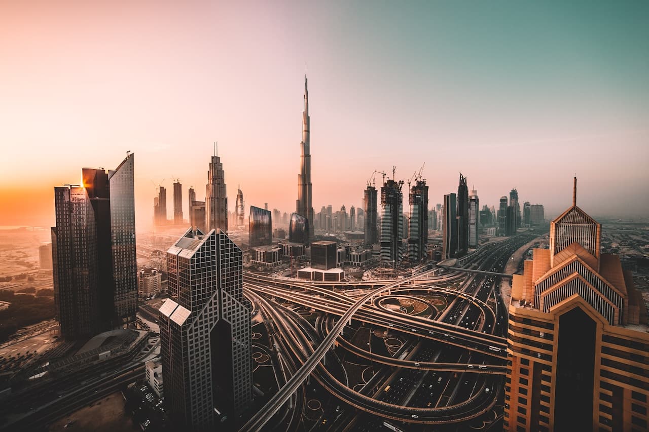 An aerial cityscape shot in warm, hazy light, featuring a sweeping highway interchange and clusters of modern skyscrapers. Curving roads and ramps form bold, flowing lines that lead toward a very tall, needle-like tower in the distance. The scene is bathed in peach and sand-colored tones, with long shadows and atmospheric haze softening the far skyline. The composition is wide and dynamic, balancing the geometry of roads with the vertical rhythm of high-rises. The overall mood feels expansive and cinematic, capturing the scale and heat of a large modern city.