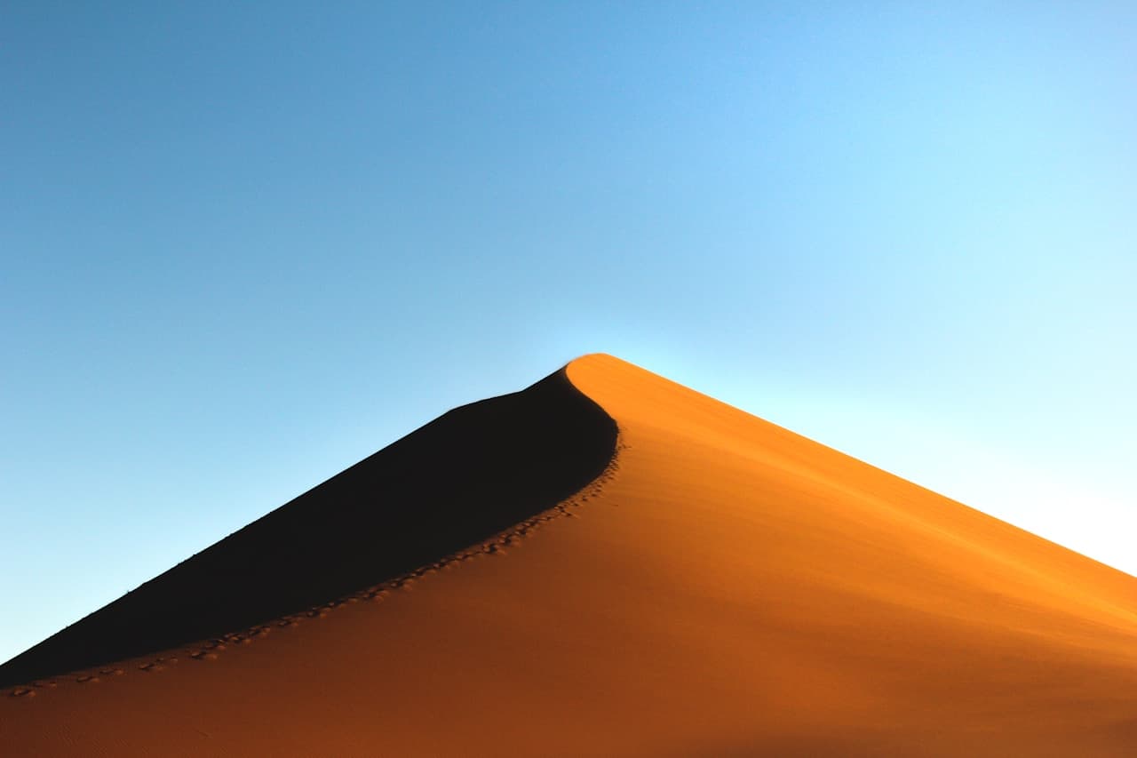 Dramatic photograph of a sharp dune peak lit by low, warm sunlight. The ridge line runs diagonally, separating a glowing orange slope from a deep, near-black shadow on the other side. A trail of footprints follows the crest, adding a small human trace without showing any people. The sky is clear and pale blue, creating strong contrast with the sand. The framing is tight and graphic, with bold shapes and clean negative space. Mood is adventurous and striking.