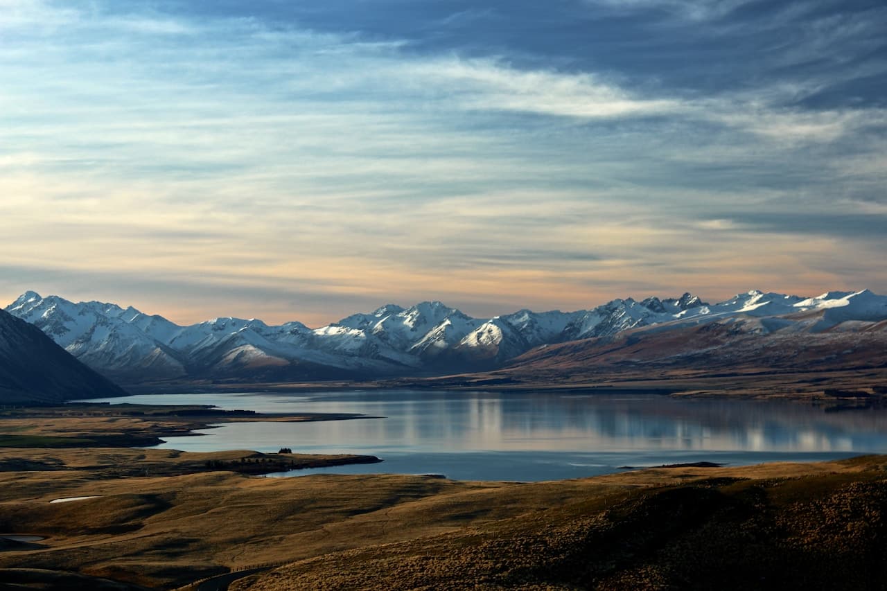 A wide panoramic photograph of a calm lake surrounded by broad, golden-brown plains and a distant line of snow-capped mountains. The water reflects the cool tones of the sky, while thin, layered clouds stretch across the frame. The composition is balanced with the horizon placed high, emphasizing the lake and landforms below. Light is soft and subdued, suggesting early morning or late afternoon, with gentle contrast and muted shadows. The mood is peaceful and spacious, conveying quiet isolation and a sense of vast open terrain.