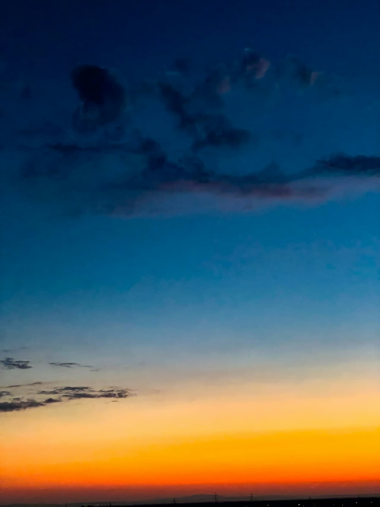 A vertical photograph focused almost entirely on the sky at dusk, with a strong gradient from deep navy and cobalt at the top to warm yellow-orange near the horizon. Dark, soft-edged clouds float across the upper portion, while the bottom edge shows a thin, distant strip of land and faint lights. The composition is minimal and graphic, emphasizing color transitions and atmospheric layers rather than specific landmarks. Light is low and fading, creating a calm, reflective mood that feels like the last moments of sunset.