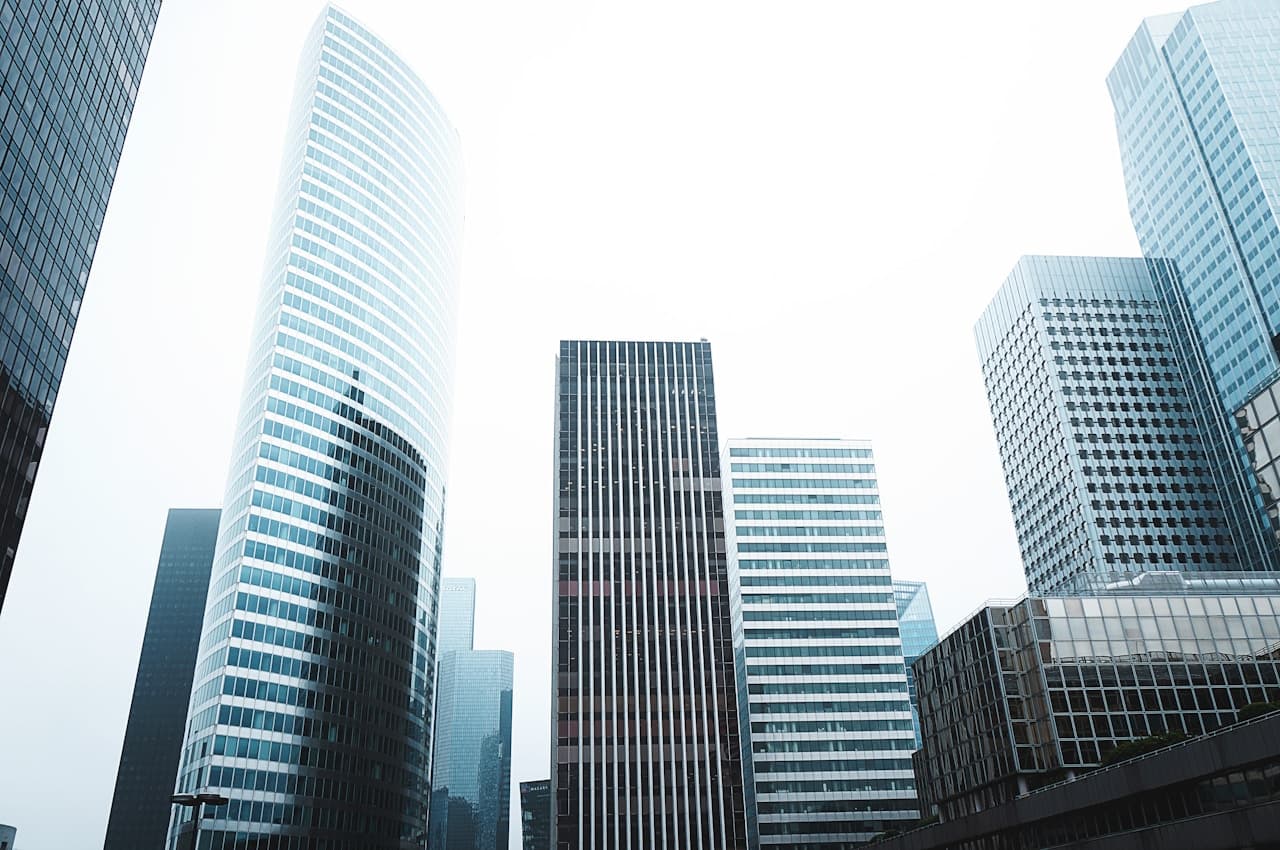 A clean, architectural photograph of modern office buildings shot from a low angle, looking up at glass and concrete facades. Vertical lines and repeating window grids create a strong geometric pattern, while the pale sky provides a minimal backdrop. Reflections in the glass add subtle blue-gray tones, and the overall palette is cool and neutral. The composition is tight and upward-focused, emphasizing height and structure rather than street activity. Lighting is soft and diffused, likely overcast, resulting in gentle contrast and a calm, contemporary mood.