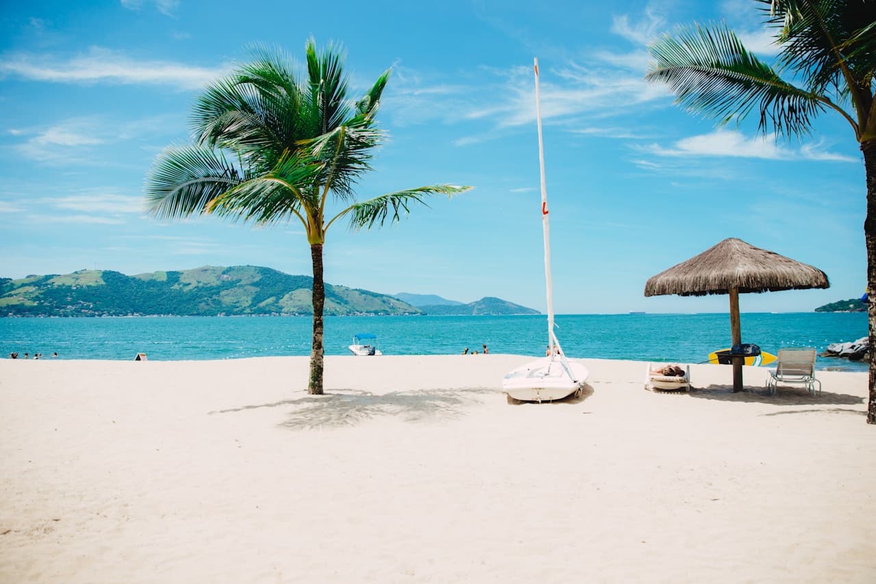 A sunny beach scene with wide white sand and a calm blue sea under a clear sky. A small sailboat rests on the sand near the center-right, and a thatched umbrella shades lounge chairs nearby, suggesting a resort-like setting. A palm tree stands to the left, with its shadow falling across the sand, while green hills or mountains line the distant horizon across the water. The photo is bright and clean, with simple horizontal layers of sand, sea, and sky. The mood is relaxed and leisurely, evoking a warm coastal holiday.