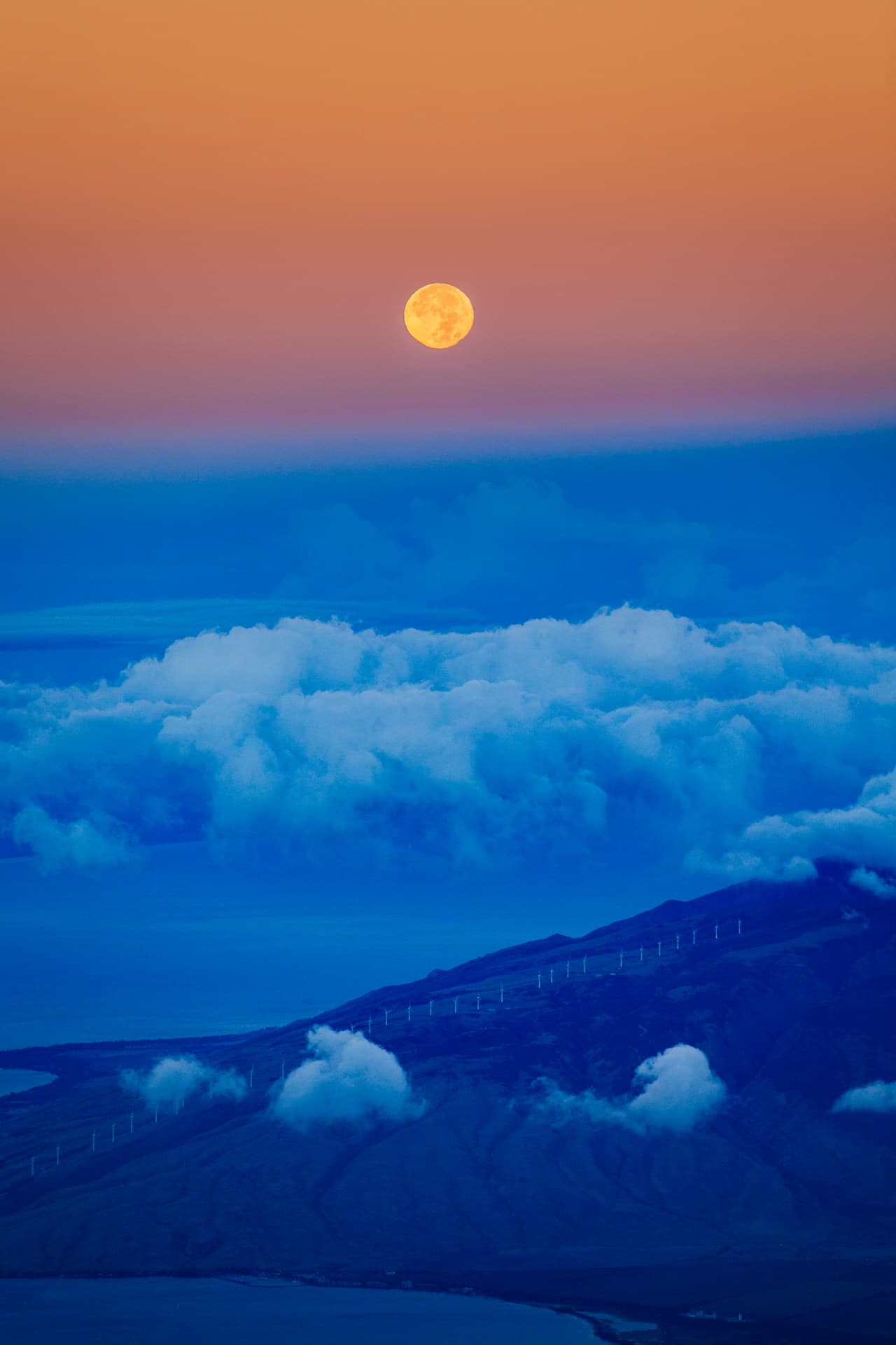 A telephoto landscape photo centered on a bright full moon floating above layered clouds. The upper sky fades from warm orange to soft mauve, while the lower half is dominated by deep blue cloud banks and a dark ridgeline. The moon acts as a clean focal point, balanced by horizontal bands of atmosphere and terrain. Light is gentle and hazy, creating a dreamy, surreal sense of scale. The mood is calm and slightly otherworldly, like a quiet moment at dusk.