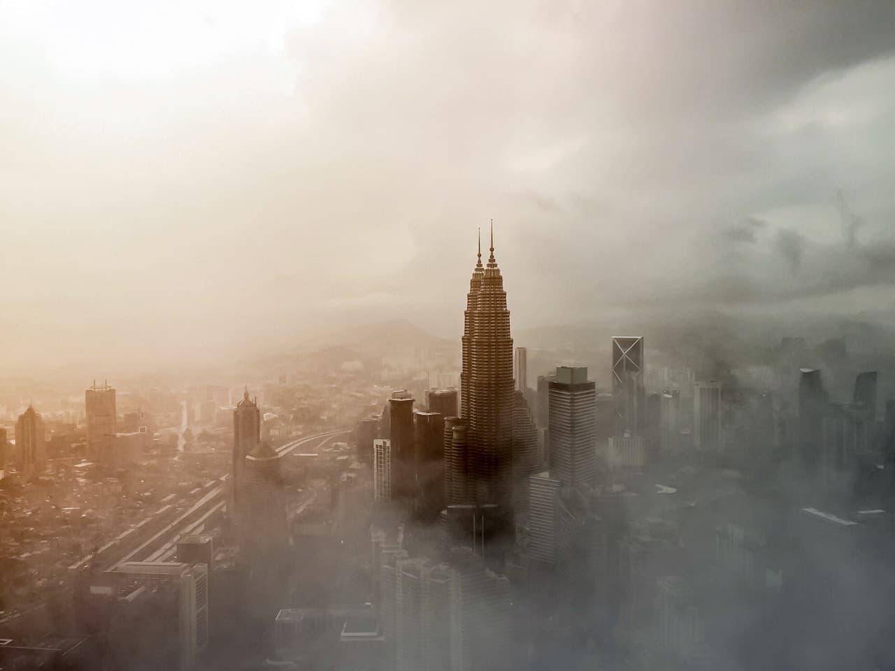 A moody skyline scene where two tall, matching towers rise above a layer of fog or low clouds, partially obscuring the city below. The towers are lit in cool whites, standing out against a gray-blue atmosphere, while a soft warm glow near the horizon suggests early morning or late afternoon light. The composition is vertical and centered, using the mist as negative space to emphasize height and symmetry. The overall look is photographic and atmospheric, with softened edges and muted contrast. The mood feels calm, distant, and slightly surreal, like the city is floating in clouds.