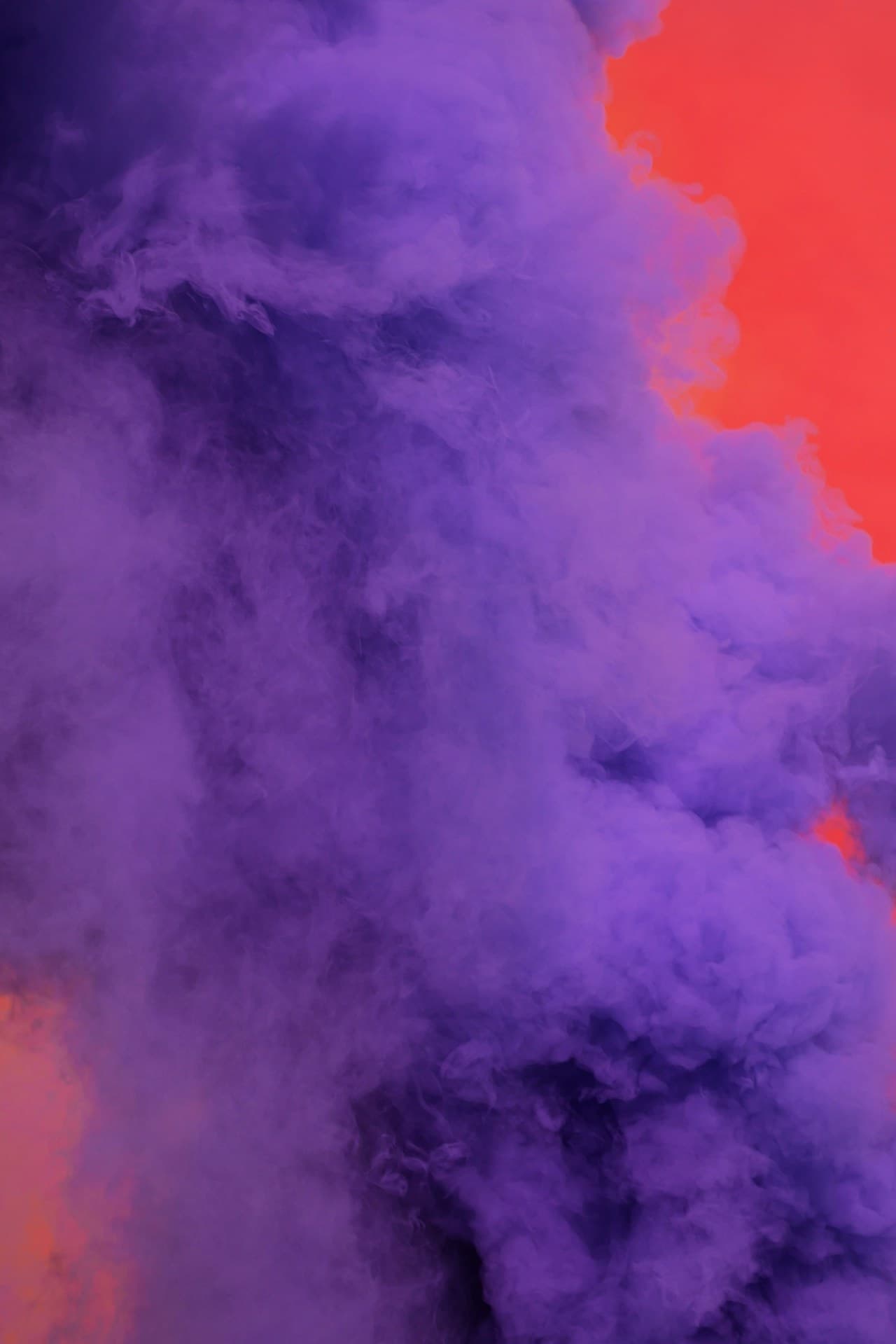 A close-up, abstract view of thick purple smoke curling and folding into itself, with warm red and orange light bleeding in from one side. The smoke forms soft, velvety layers and rounded contours, creating a painterly, almost cosmic feel. The background is dark and saturated, helping the violet tones stand out while the warm highlights add drama and depth. The composition is tight and immersive, focused on texture and color gradients rather than a defined subject, evoking a moody, atmospheric vibe.