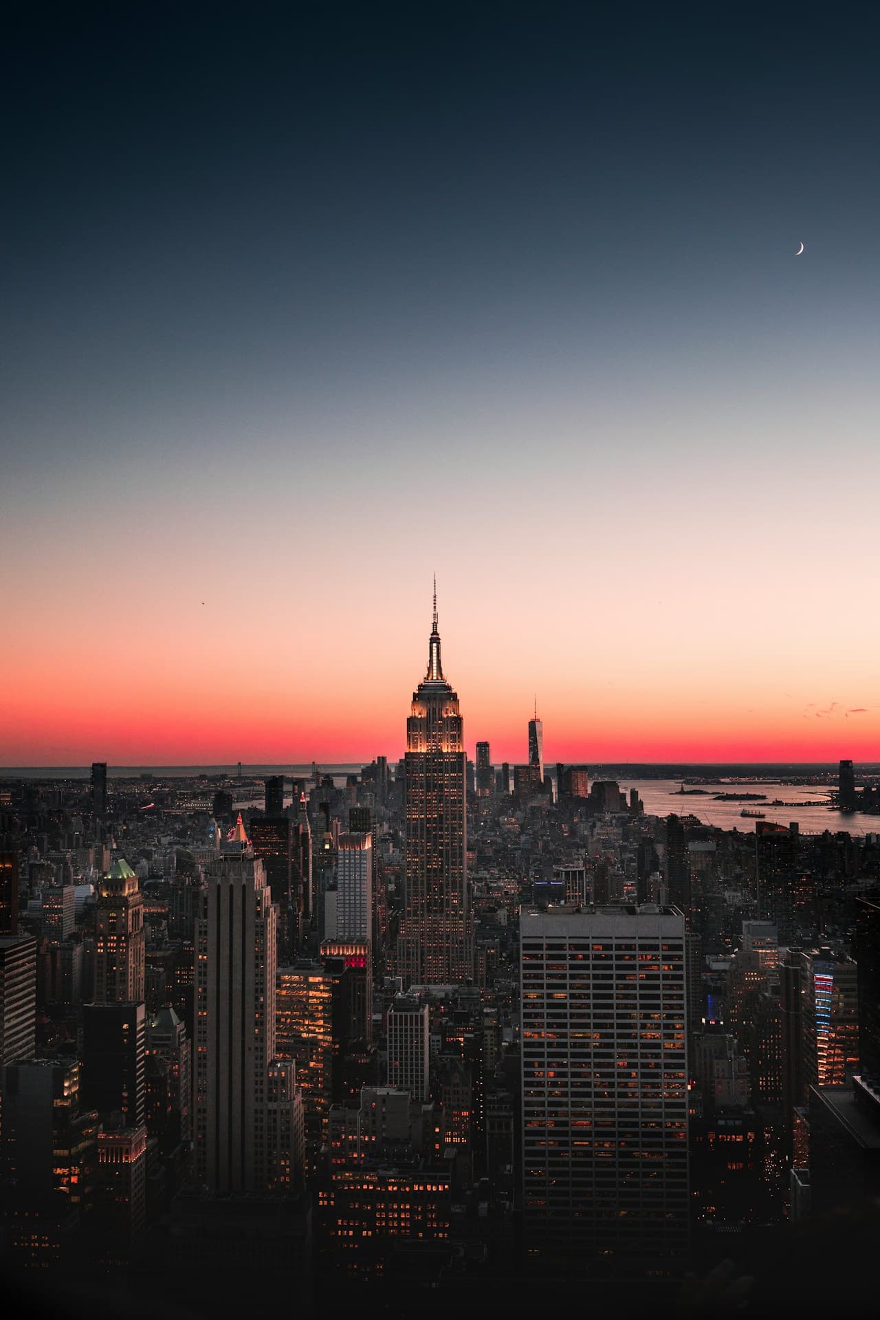 A twilight city skyline with the Empire State Building rising prominently near the center, surrounded by darker high-rises and a band of warm color on the horizon. The sky transitions from pale peach near sunset to deep blue above, while scattered city lights begin to glow. The composition is a wide, layered panorama that highlights the building's iconic silhouette without focusing on street-level detail. Low-light conditions create strong contrast and a calm, cinematic atmosphere, with haze softening distant structures. The mood feels quiet and dramatic, capturing the city as it shifts from day to night.