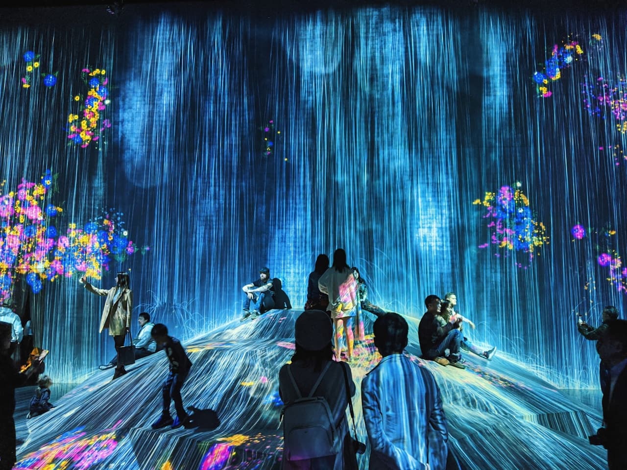 A photograph of an immersive digital art installation where tall, vertical blue light patterns resemble cascading water across the walls. Bright, flower-like bursts of color and glowing particles add contrast to the dark room. Several visitors appear as small silhouettes in the foreground, some standing and some seated on a raised, mound-like platform, emphasizing scale. The composition is wide and enveloping, with the luminous projection acting as the main subject. Low, dramatic lighting creates a futuristic atmosphere. The mood is awe-filled, calm, and otherworldly.