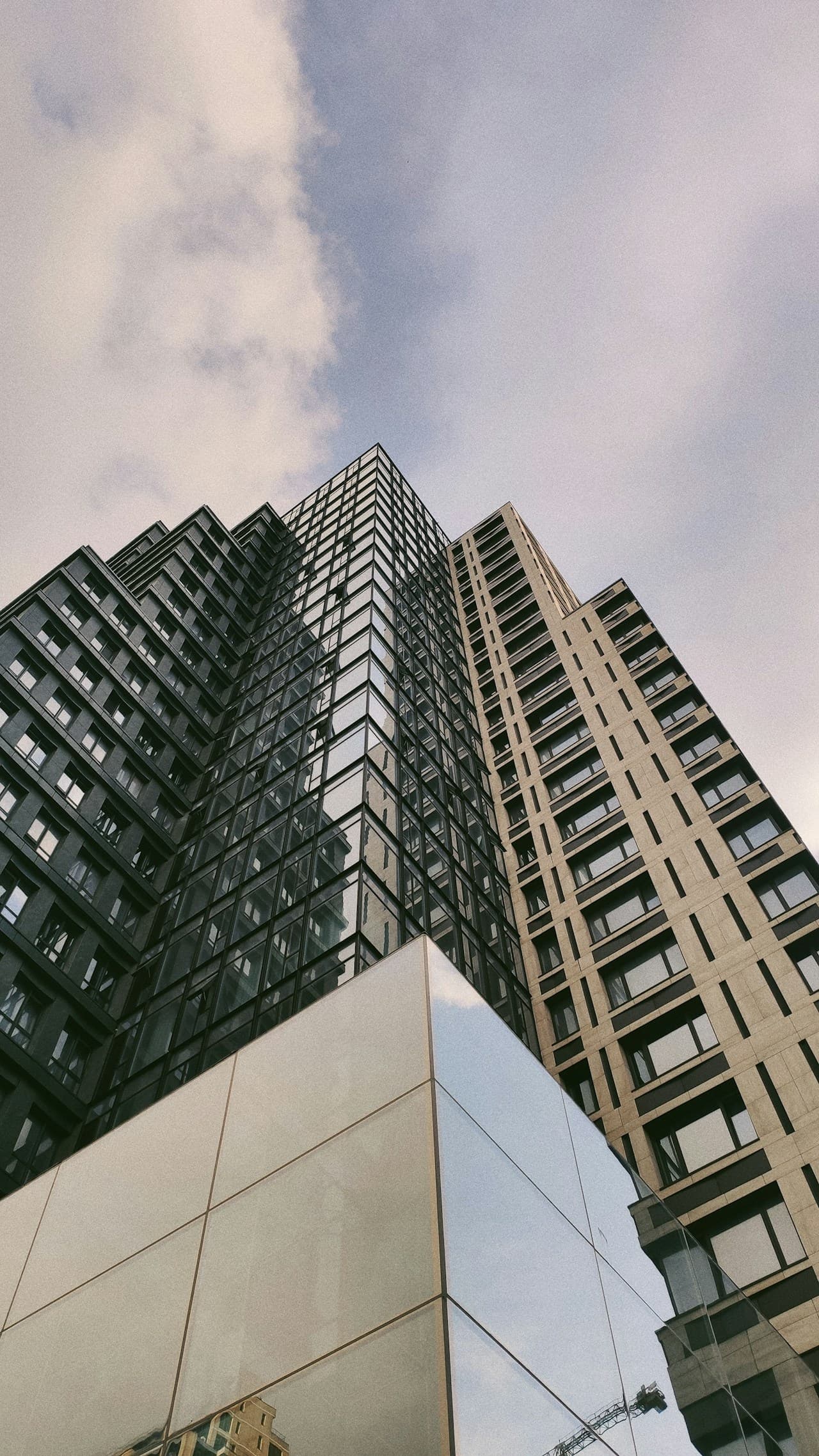 Low-angle architectural photograph looking up between modern high-rise buildings. Glass and concrete facades form strong vertical lines that converge toward the center, with a pale, cloudy sky filling most of the negative space. Windows and structural grids create repeating patterns, while the perspective exaggerates height and scale. Light is soft and overcast, producing muted tones and minimal glare. The mood is urban and clean, emphasizing geometry rather than street-level detail.