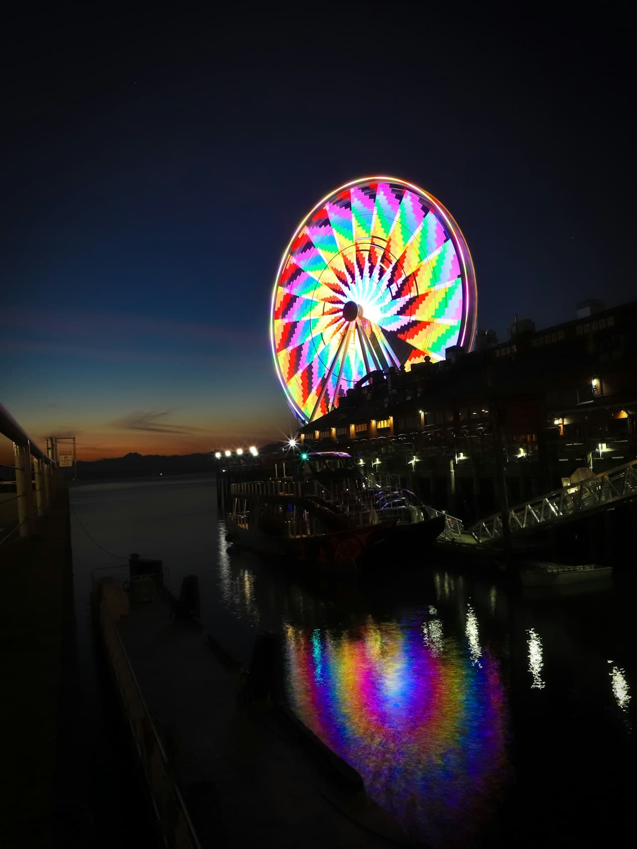 A night waterfront photograph featuring a large ferris wheel glowing with multicolored LED lights. The wheel sits beside a pier lined with low buildings and small lamps, while the calm water below mirrors the rainbow pattern in shimmering reflections. The sky is a deep gradient from dusk to night, leaving generous negative space around the bright circle. The image feels like a long exposure, with crisp light shapes against dark surroundings and a lively, festive mood.
