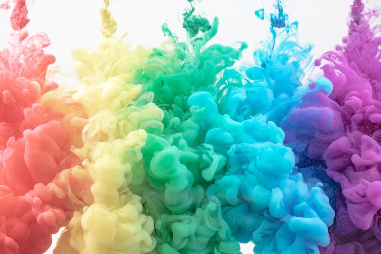 A vivid abstract photograph of colored ink or smoke clouds swirling through a clean white background. Multiple plumes overlap in a smooth gradient—reds and oranges near the bottom, transitioning through yellow and green into cyan, blue, and hints of purple at the top. The shapes are soft and rounded, with wispy tendrils that suggest fluid motion frozen mid-bloom. The composition feels centered and layered, like a painterly spectrum suspended in air. Bright, even lighting keeps it crisp and cheerful, with an energetic, playful mood.