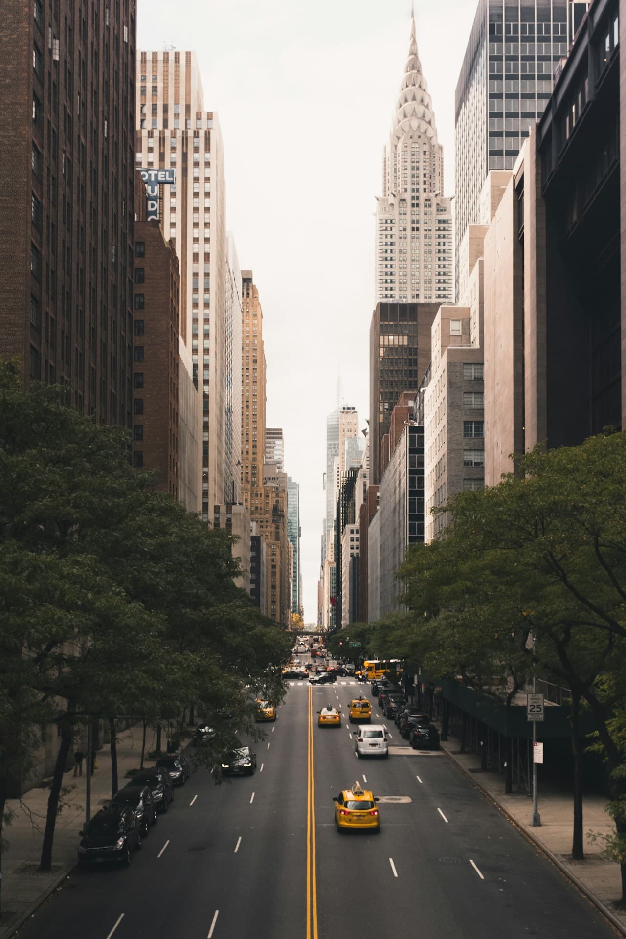 A classic city street view framed by tall buildings and lined with trees, with an Art Deco-style skyscraper spire visible in the distance. The shot is taken from street level along the centerline, creating strong perspective and depth. Traffic and taxis appear as small moving elements, while pedestrians are secondary and not individually discernible. The lighting is bright but slightly diffuse, suggesting a mild or overcast day. The palette is mostly neutral—stone facades, asphalt, and muted greens—punctuated by occasional yellow vehicles. The mood feels lively and metropolitan, with a timeless urban rhythm.