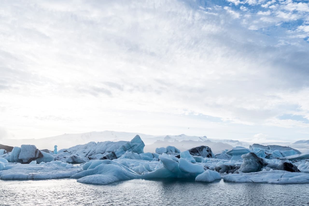 A cold, high-key landscape of floating ice and calm water under a bright, overcast sky. Chunky blue-white ice formations stretch across the midground, with smooth water in the foreground reflecting the pale light. Distant, faint landforms sit on the horizon, partially softened by haze. The composition is a wide shot with a low viewpoint, emphasizing the texture and sculptural shapes of the ice. Colors are muted and cool, and the soft lighting gives the scene a quiet, almost ethereal atmosphere.