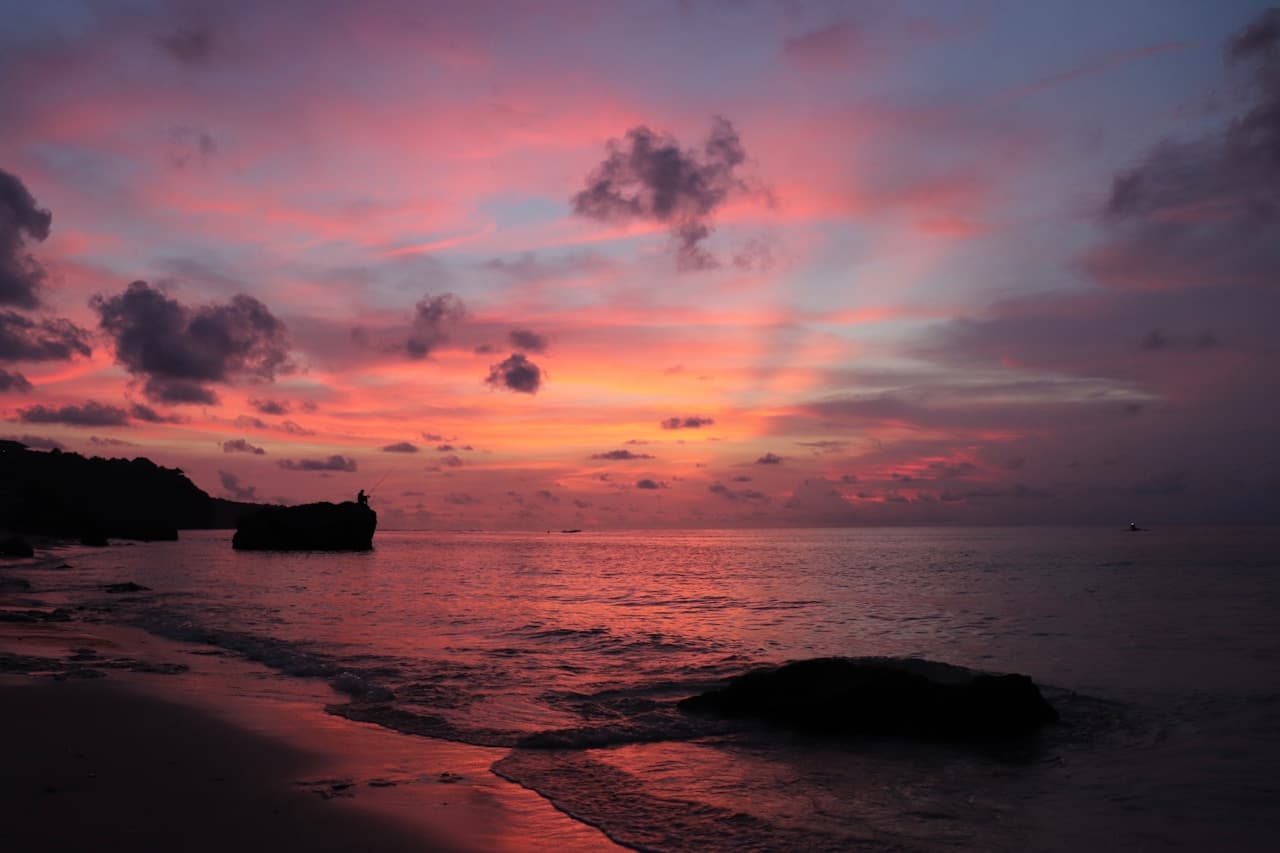 A sunset seascape with a gently curving shoreline in the foreground and a calm, open horizon beyond. The sky is painted in layers of pink, coral, and soft purple, with scattered clouds catching the last light. Dark rock formations and a distant headland appear as silhouettes, adding contrast and depth. Wet sand near the waterline reflects the warm colors, creating a subtle mirror effect. Shot as a wide landscape, the scene feels peaceful and romantic, with soft, low light and a tranquil evening atmosphere.