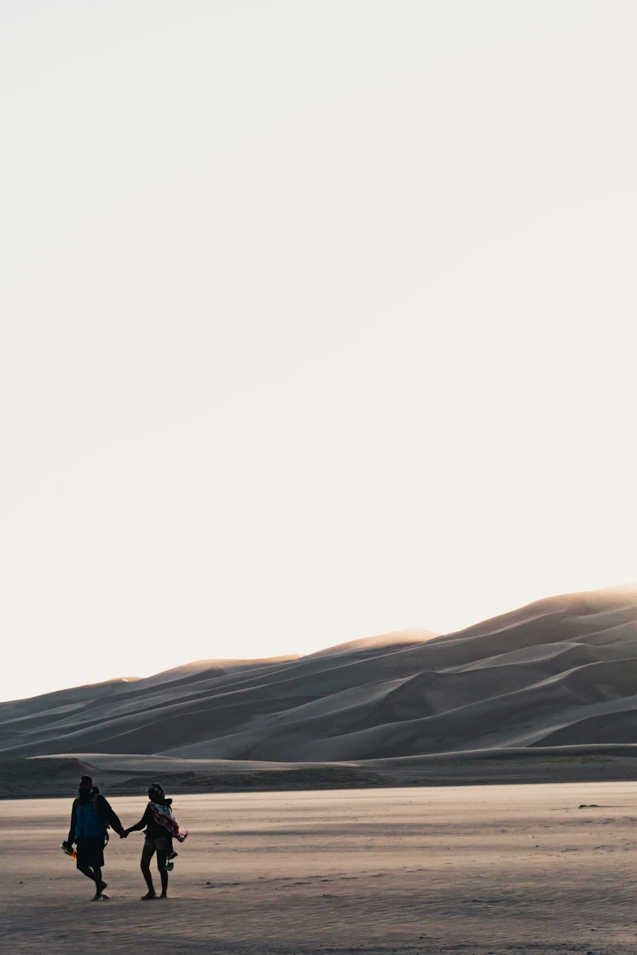 Minimal desert scene with an enormous blank sky occupying most of the frame. Two small figures walk side by side across a flat sandy foreground, while distant dunes rise in soft layers along the horizon. The light is gentle and hazy, muting the colors into grays and pale beiges. The composition feels cinematic and spacious, with the human silhouettes anchored low in the frame. No buildings or distinct landmarks are visible. Mood is quiet, intimate, and expansive.