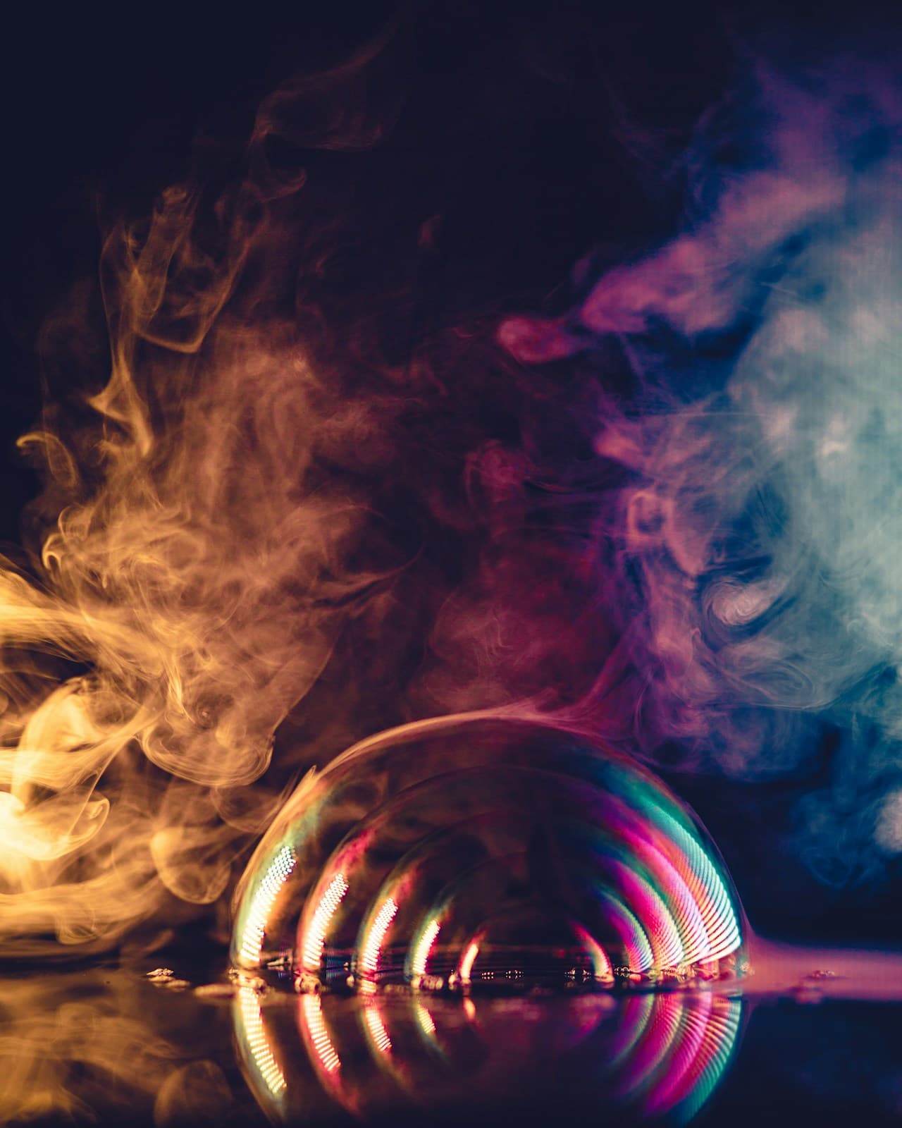 A dark, surreal still-life featuring an iridescent soap bubble resting on a reflective surface, surrounded by drifting smoke. Colored plumes—yellow on the left, magenta in the middle, and blue on the right—frame the bubble and echo in the glossy reflections below. The scene is shot in low light with a studio-like feel, using deep blacks and selective highlights to emphasize the bubble's curved sheen and the soft, foggy texture of the smoke. The composition is centered and theatrical, creating a mysterious, dreamlike mood.