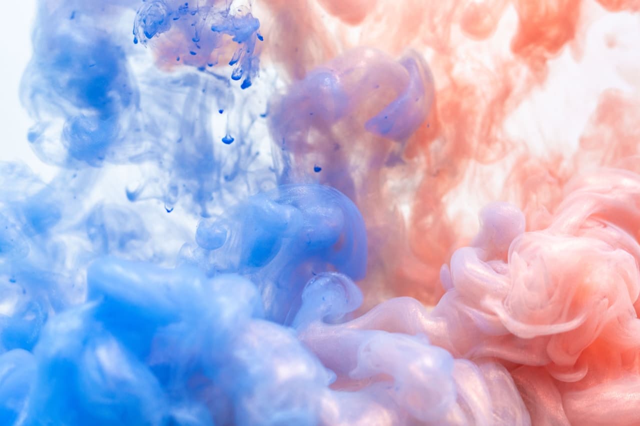 A soft, high-key abstract image of pastel smoke or ink clouds mixing in a white, airy space. Cool powder-blue dominates one side while warm peach and coral tones billow alongside it, blending into gentle purples where the colors overlap. The forms are rounded and cloud-like, with thin wisps dissolving into the background. The composition is balanced and minimal, emphasizing color harmony and fluid texture rather than sharp detail. Even, diffused lighting creates a clean, modern look and a calm, uplifting mood.