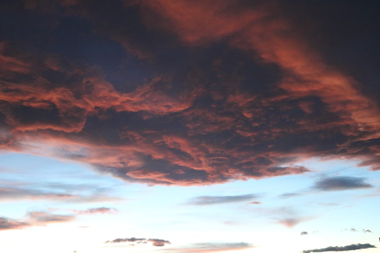 A dramatic sky photograph dominated by a heavy ceiling of dark storm clouds. The underside of the cloud mass glows with warm red and orange tones, as if lit by a low sun, while a strip of pale blue sky remains near the horizon. The contrast between the fiery highlights and the charcoal shadows creates a tense, cinematic atmosphere. The framing is upward-looking and wide, filling the scene with texture and swirling cloud shapes. Overall mood is moody and imminent, like weather changing at dusk.