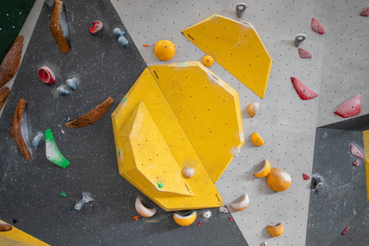 Indoor climbing wall photographed straight on, featuring large geometric yellow volumes and scattered climbing holds in red, orange, and green. The wall surface is light gray and textured, with bolt holes and panel seams visible. The bright yellow structures dominate the frame, creating bold shapes and shadows that add depth. Composition is tight and graphic, focusing on pattern and color rather than people. Lighting is even and indoor-soft, giving the scene a playful, sporty mood.