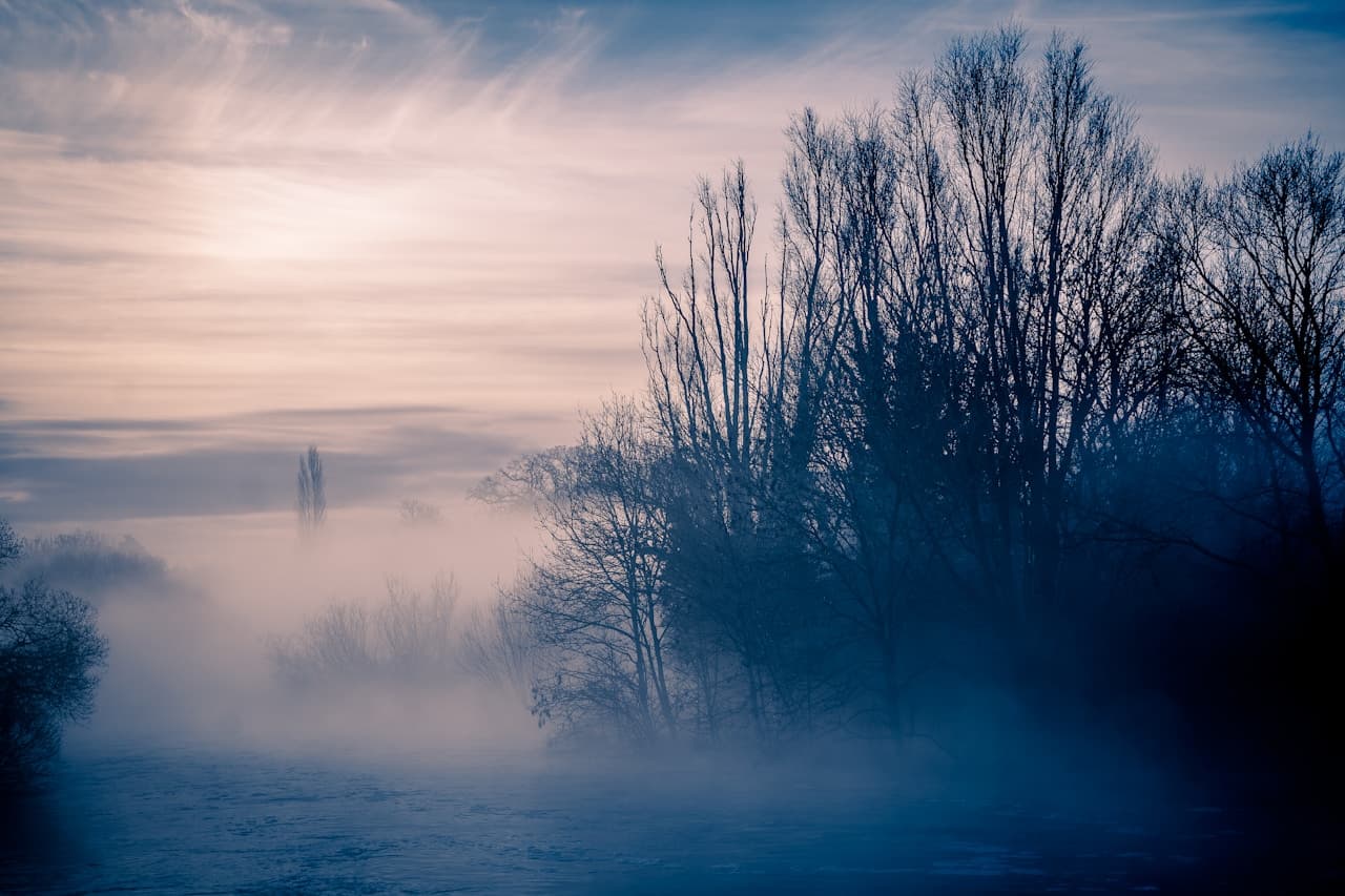 A minimal, moody landscape at dawn with a winding river or wetland channel cutting through flat fields. Bare trees and sparse vegetation sit as silhouettes against a cool blue haze, while thin fog softens the horizon and reduces contrast. The composition is wide and open, with the water forming gentle curves that lead the eye into the distance. Light is soft and diffused, suggesting early morning calm before full daylight. The overall mood is quiet, contemplative, and slightly melancholic, emphasizing stillness, empty space, and muted color.