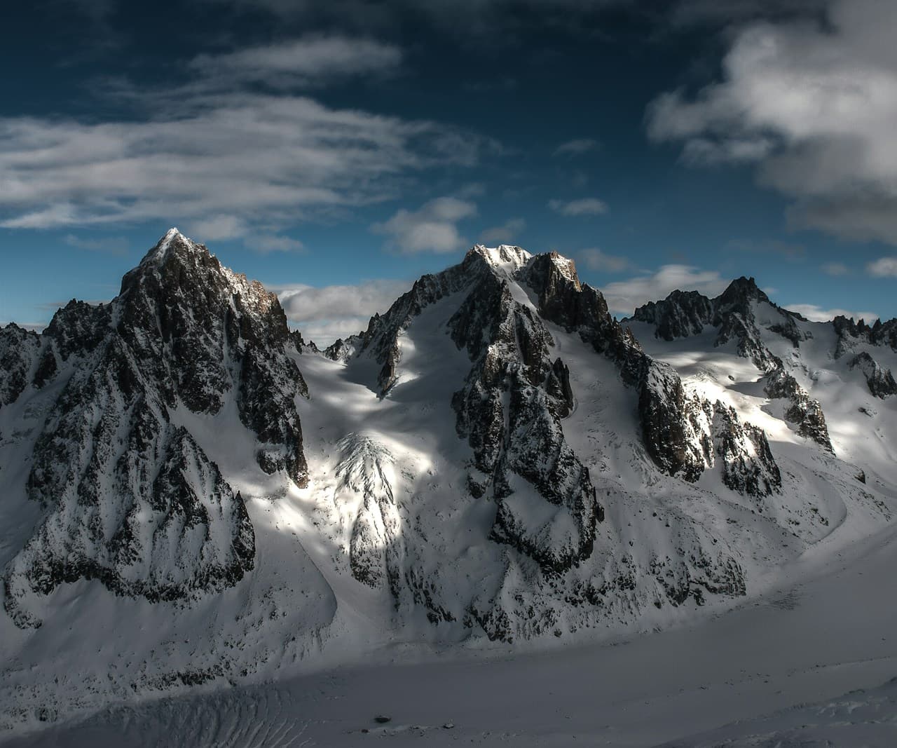 A dramatic mountain landscape featuring jagged, rocky peaks covered in snow beneath a deep blue sky. Sunlight breaks through scattered clouds, creating bright highlights on the snowfields and strong shadows that carve out the ridges. The composition is wide and layered, with multiple spires receding into the distance and a broad, frozen basin in the foreground. The scene feels crisp and cold, with high-altitude clarity and a bold, adventurous mood.