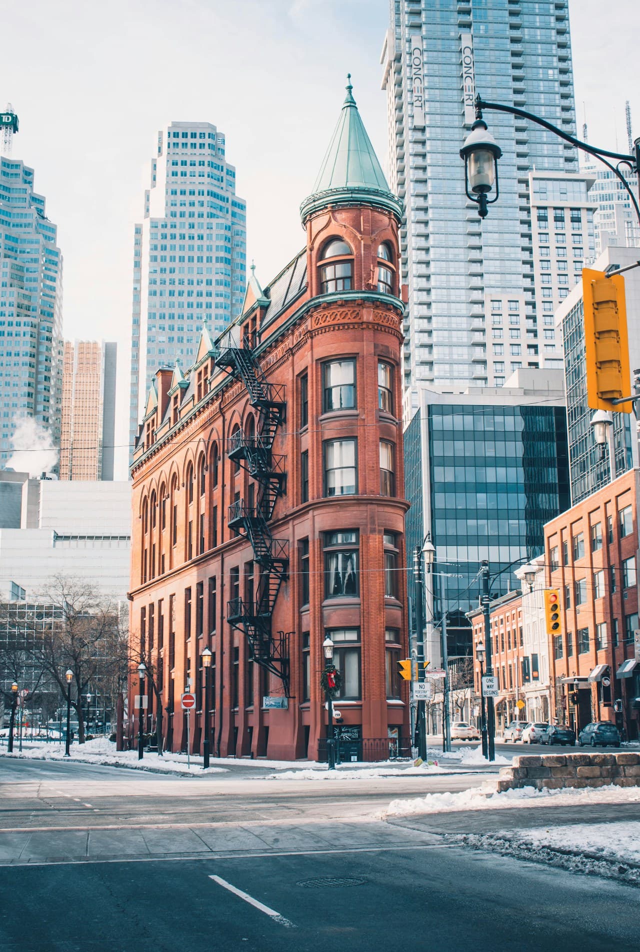 A street corner scene dominated by a tall, red-brick building with a flatiron-like angle, photographed in cold daylight. Snow lines the sidewalks and street edges, while the intersection and surrounding buildings frame the architecture. The brick facade adds warmth against the otherwise cool palette of pale sky, gray pavement, and muted glass towers in the background. The composition places the building as a strong central anchor, with streets leading away on both sides. Lighting is soft and overcast, giving the scene a quiet, crisp mood—urban and slightly nostalgic.