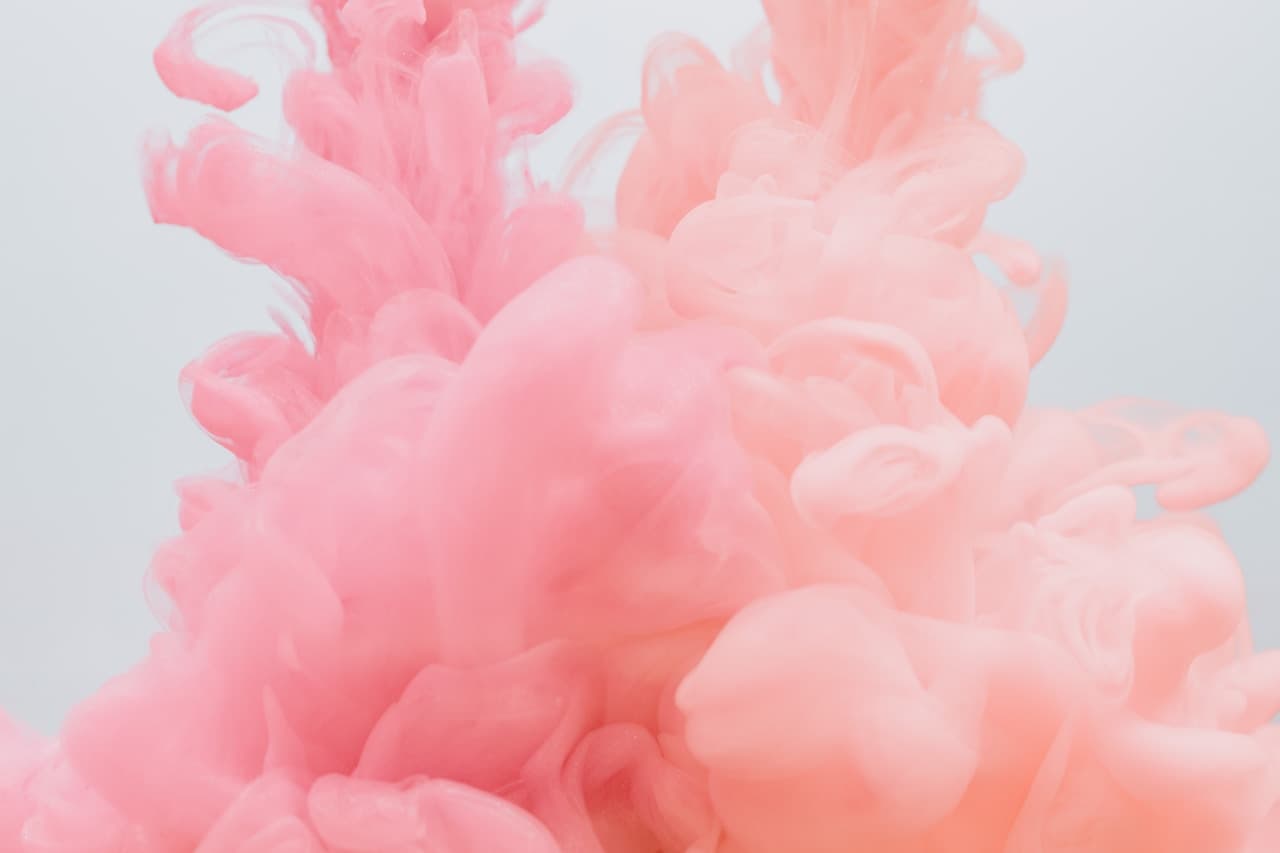A minimal, airy composition showing a soft pink smoke or ink cloud suspended against a pale gray-white background. The plume spreads diagonally with delicate, translucent folds and feathered edges, creating a gentle sense of motion. With no hard lines or objects, the image feels like a studio-style abstract photograph focused on texture and subtle gradients. The light is diffuse and even, producing a clean, high-key look. Overall mood is calm and dreamy, like a pastel watercolor floating in space.