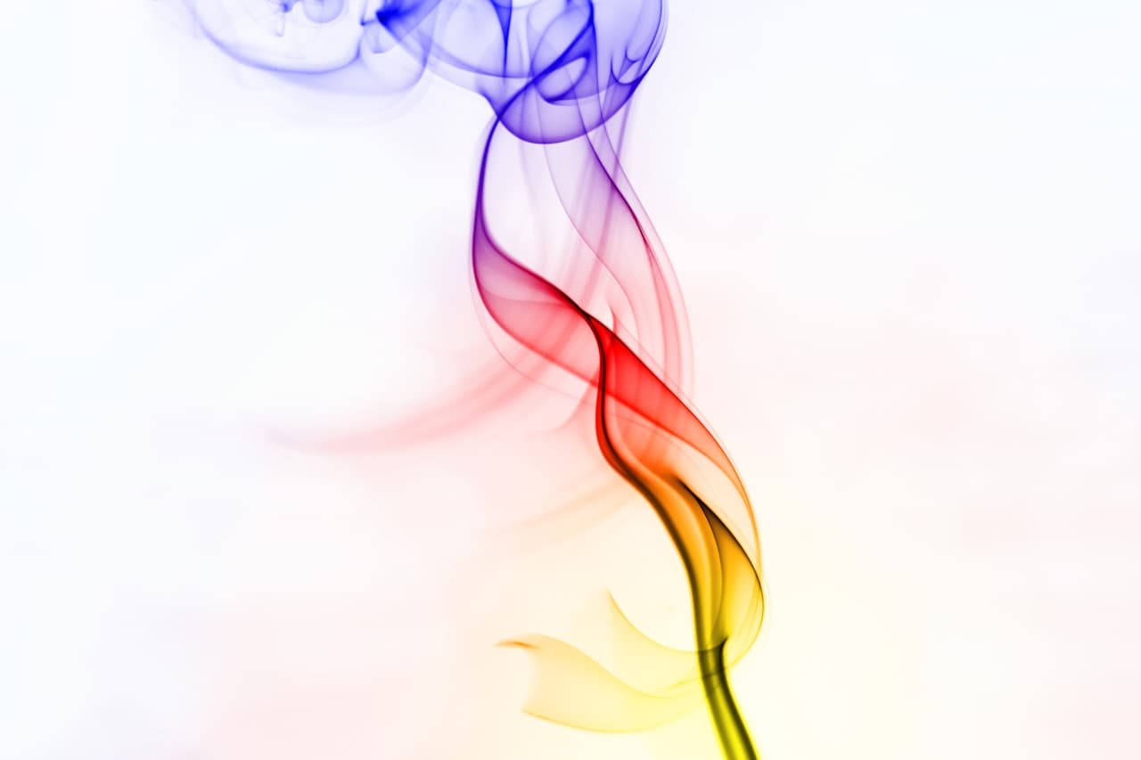A minimalist abstract photograph showing a thin plume of smoke or ink rising through a bright white background. The column transitions from yellow at the base to warm red and orange, then into deep purple and blue as it curls at the top. Delicate wisps peel away from the main stream, creating fine, feathery textures and a sense of upward motion. The composition is centered and elegant, with ample negative space and soft, even lighting. The mood feels clean and modern, like a single brushstroke of color suspended in air.
