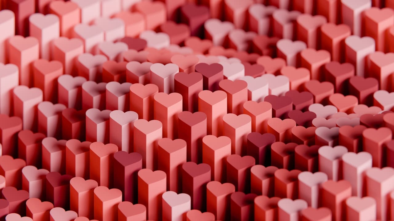 A playful 3D pattern of heart-shaped pillars packed tightly together. The hearts rise to different heights and range from deep red to rose pink and pale blush, with soft shadows that add depth and a gentle focus toward the center. The camera angle is slightly elevated, letting the repeating shapes fill the frame edge to edge like a decorative texture. Lighting is soft and even, giving the hearts a smooth, matte look. The overall mood is warm and romantic, with a candy-like color palette.