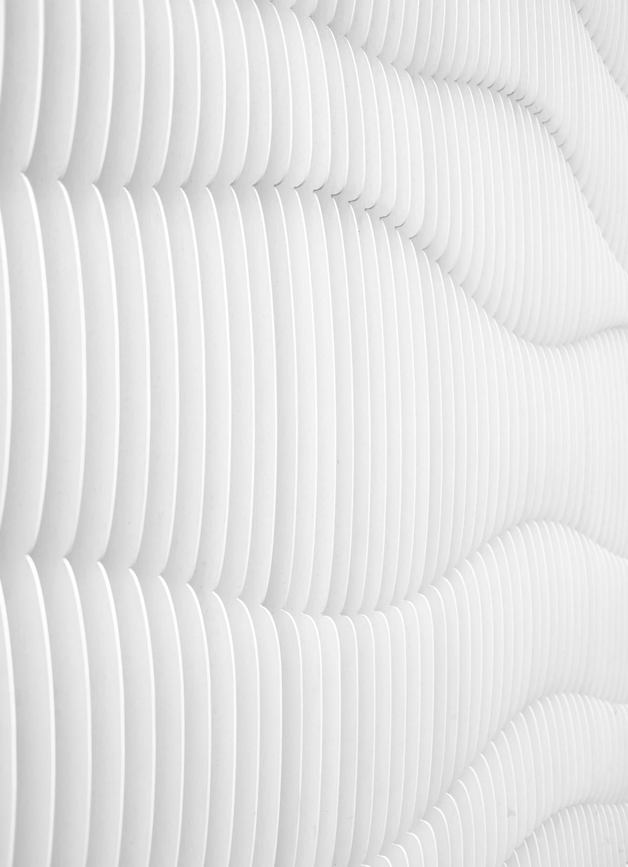 Minimal architectural texture of a white ribbed surface made of repeating vertical grooves. The ridges curve subtly, creating a wave-like rhythm across the frame. Soft side lighting casts delicate shadows into the recesses, revealing depth without strong contrast. There are no other objects or colors—just variations of white and light gray—so the image reads as an abstract pattern. The framing is close and immersive, with the lines extending beyond the edges. Mood is calm, clean, and modern.