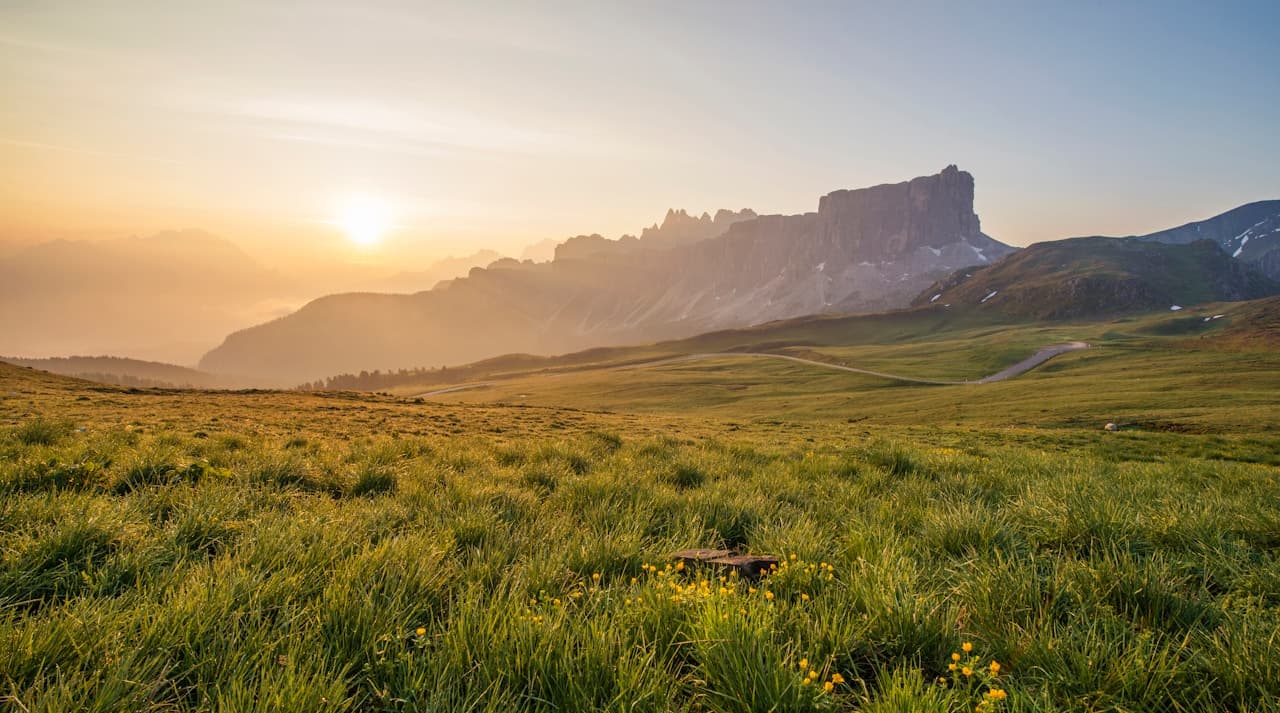 A golden-hour landscape of an alpine meadow stretching toward a rugged mountain ridge. The sun sits low near the horizon, casting warm light across the grass and creating a hazy glow in the distant valleys. Subtle trails and gentle curves in the terrain guide the eye toward the peaks, while the sky transitions from warm amber near the sun to cooler blue above. The composition is wide and open, emphasizing space and light. The mood feels hopeful and serene, like an early morning hike with crisp air and soft, radiant color.