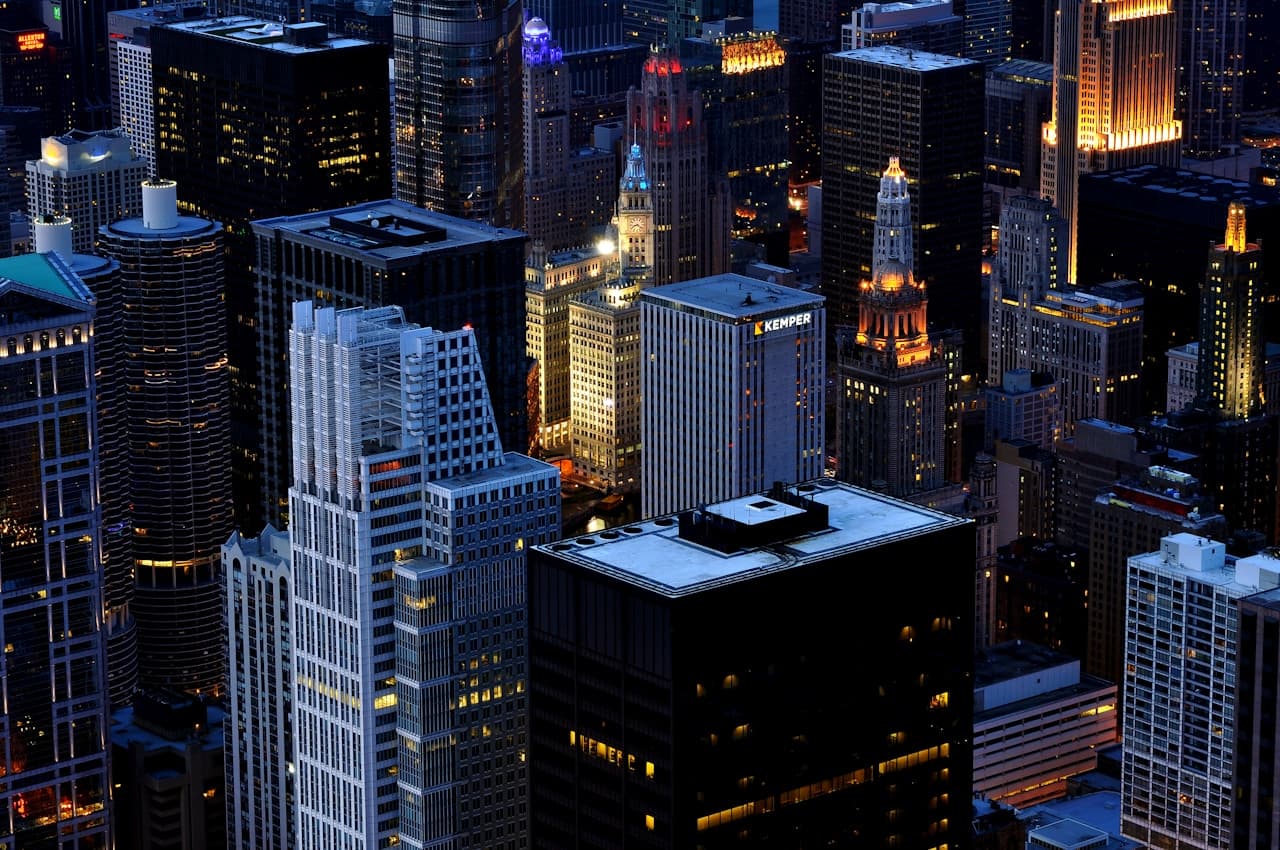 A night view of a downtown cluster of skyscrapers, photographed from an elevated vantage point. Windows and streetlights glow in warm amber and cool white, creating a glittering pattern against dark building silhouettes. The scene leans cool overall, with deep navy shadows and occasional blue highlights, suggesting late evening conditions. The composition is tightly framed around the high-rises, emphasizing vertical lines, repeating grids, and the density of the urban core. Low-light sharpness and contrast give it a crisp, modern feel. The mood is vibrant and metropolitan, quietly intense.