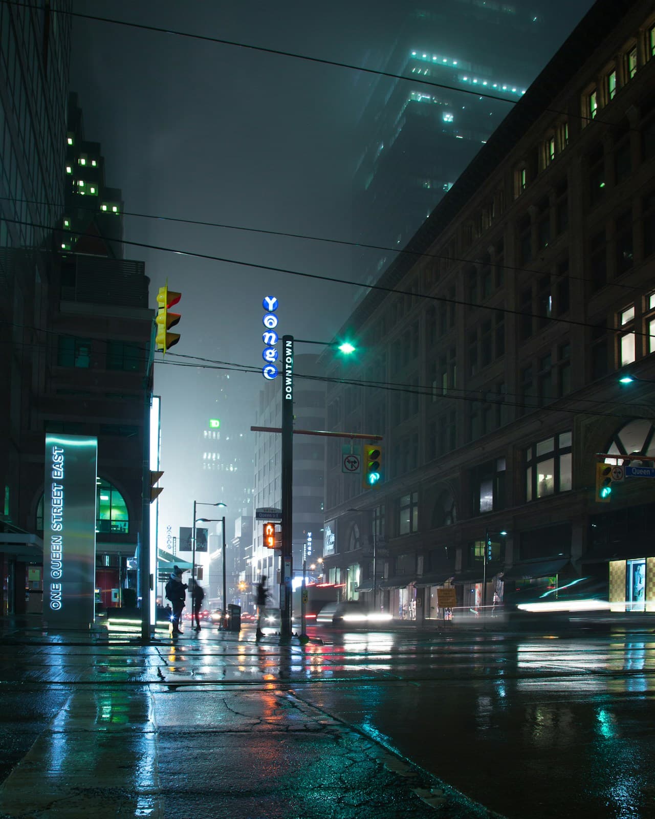 A rainy night street scene with heavy atmosphere: wet pavement, mist, and glowing signage reflecting off the road. A bright sign and streetlights create pools of warm light, while cool teal and greenish tones dominate the shadows and building facades. Small figures appear under umbrellas or in silhouette, but remain indistinct and secondary to the environment. The camera frames the street frontally, letting reflections and haze build depth. The scene is low-lit and cinematic, with a moody, urban noir feel—quiet, damp, and luminous with scattered color.