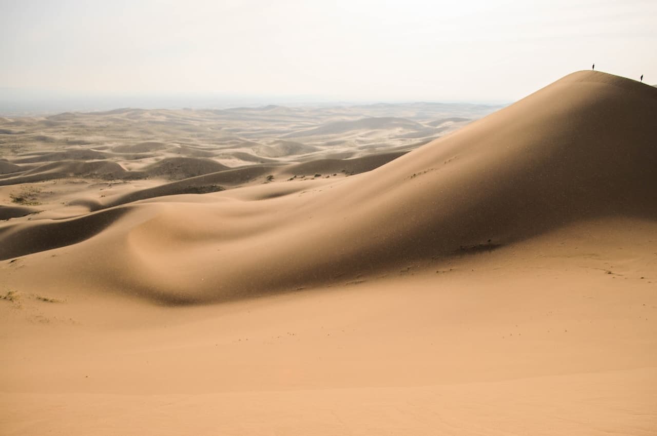 An expansive dune landscape seen from a high vantage point, with a steep, smooth sand slope rising on the right and a sea of smaller dunes fading into a hazy horizon. Tiny human figures stand near the crest, providing scale against the immense terrain. The light is soft and slightly diffused, washing the scene in warm beige tones and gentle shadows. The composition uses sweeping curves and negative space, creating a tranquil, airy atmosphere that emphasizes distance, wind, and the desert's vast openness.
