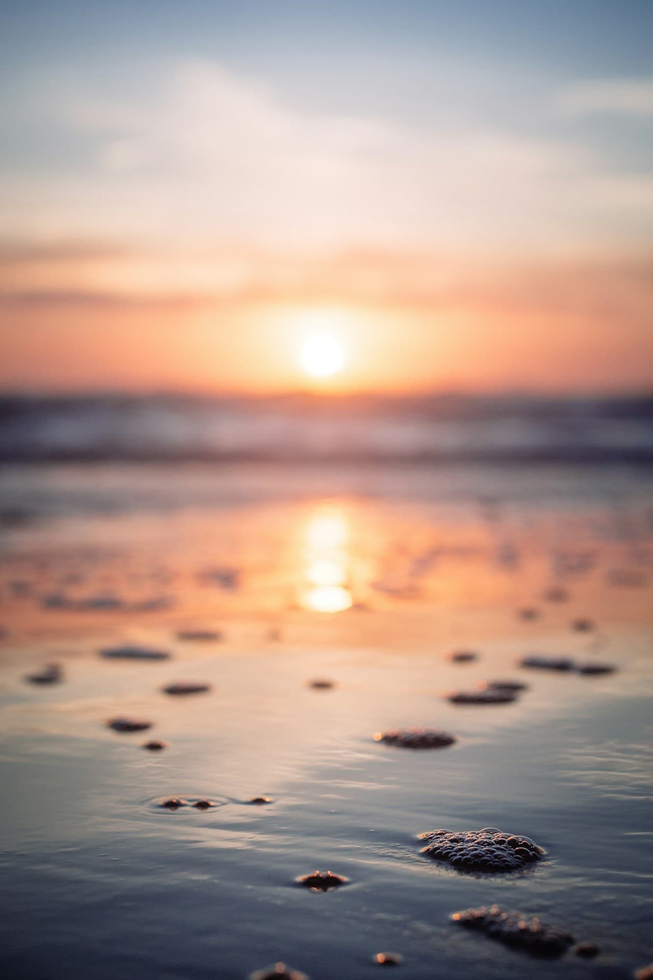 A dreamy, shallow-focus photograph of a beach at sunset, intentionally blurred to emphasize color and atmosphere rather than detail. The sun appears as a soft glowing orb near the horizon, and its reflection stretches down the center of the frame across wet sand and shallow water. Small bubbles and ripples in the foreground become gentle shapes, while the ocean and sky dissolve into bands of peach, pink, and cool blue. The composition is minimal and abstract, with soft light and a tranquil, meditative mood.