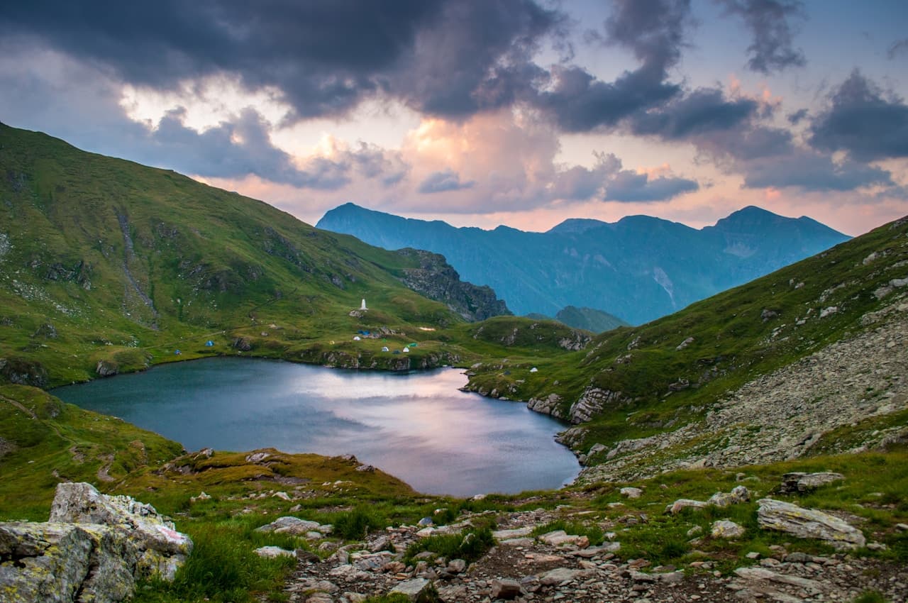 A wide mountain landscape centered on a small, still lake nestled between green slopes. Rocky ground and a faint trail occupy the foreground, leading the eye toward the water's reflective surface. Beyond, layered mountain silhouettes fade into cool blue tones, while thick clouds overhead catch hints of pink and warm light, suggesting late afternoon or early evening. Tiny tents or small structures dot the far shore, adding scale to the scene. The lighting is soft and atmospheric, creating a calm, outdoorsy mood that feels like a quiet campsite in the highlands.