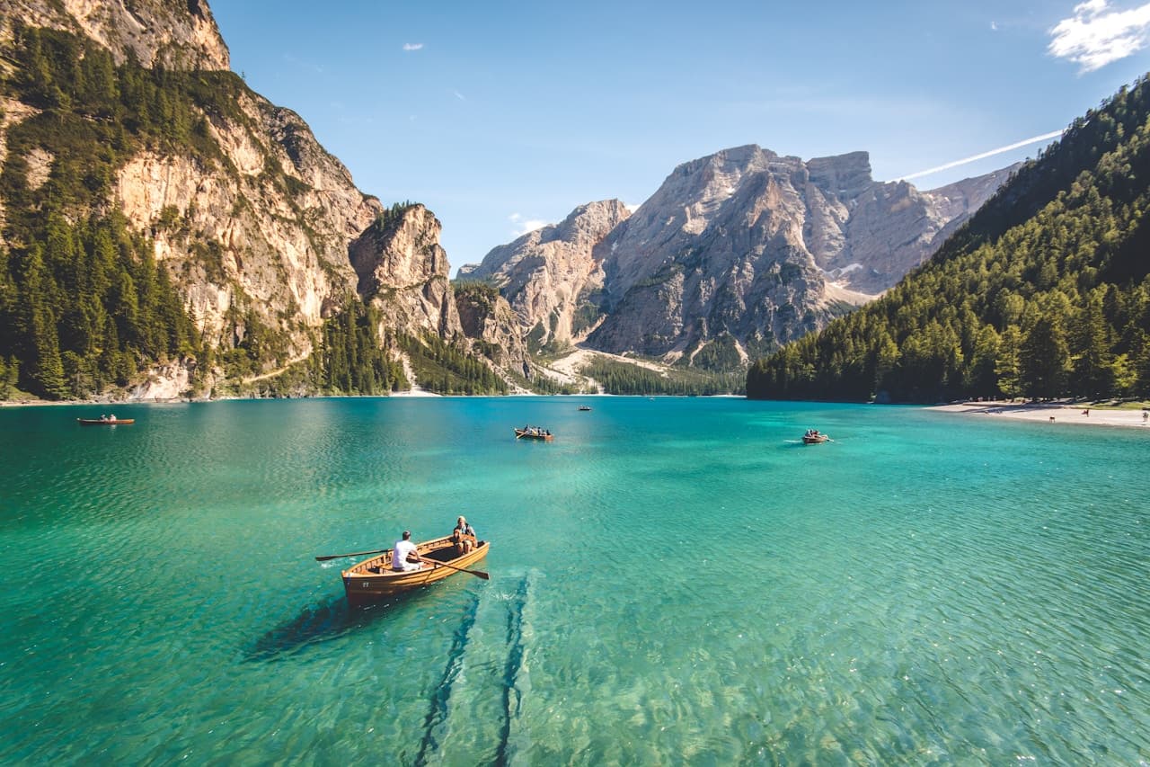 A bright mountain-lake photograph with turquoise water in the foreground and forested slopes rising toward rugged peaks. Several small rowboats float on the lake, with tiny figures visible inside, adding scale and a sense of activity. The composition is a wide shot that layers water, shoreline trees, and mountains under a clear sky. Daylight is crisp and high-key, with reflections on the water and soft shadows on the hillsides. The mood is fresh and inviting, suggesting a clear, cool day outdoors and a leisurely moment on the water.