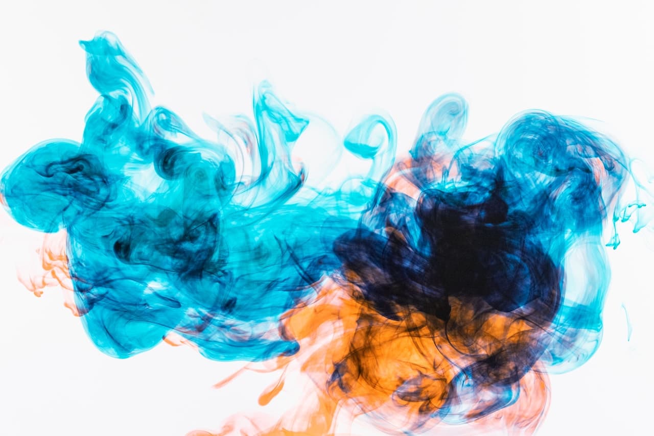 An abstract ink-in-water style image with a dense teal-blue cloud spreading across a bright white background. A dark navy core sits near the center, while small bursts of orange and rust-colored pigment flare outward, creating sharp contrast and a sense of turbulence. The composition is horizontal and expansive, with soft, rounded forms and thin wisps that fade into the white negative space. Lighting is even and clean, emphasizing subtle gradients and fine textures. The mood is energetic yet modern, like a controlled fluid experiment captured mid-bloom.