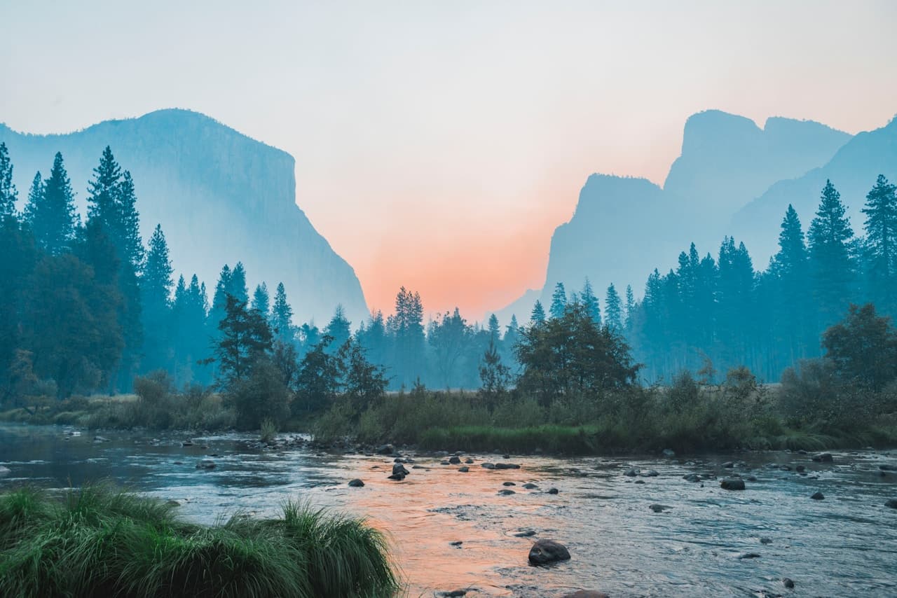 A serene landscape photograph of a river flowing through a forested valley, framed by tall pines and distant granite cliffs. Morning mist hangs in the air, softening the background and creating layered depth. Warm sunlight touches parts of the trees and rock faces, while the river reflects cool blues and silvery highlights. The composition uses the winding water as a leading line into the scene, captured in a wide, cinematic view. Lighting is gentle and atmospheric. The mood is quiet and contemplative, like an early morning moment before the day fully begins.