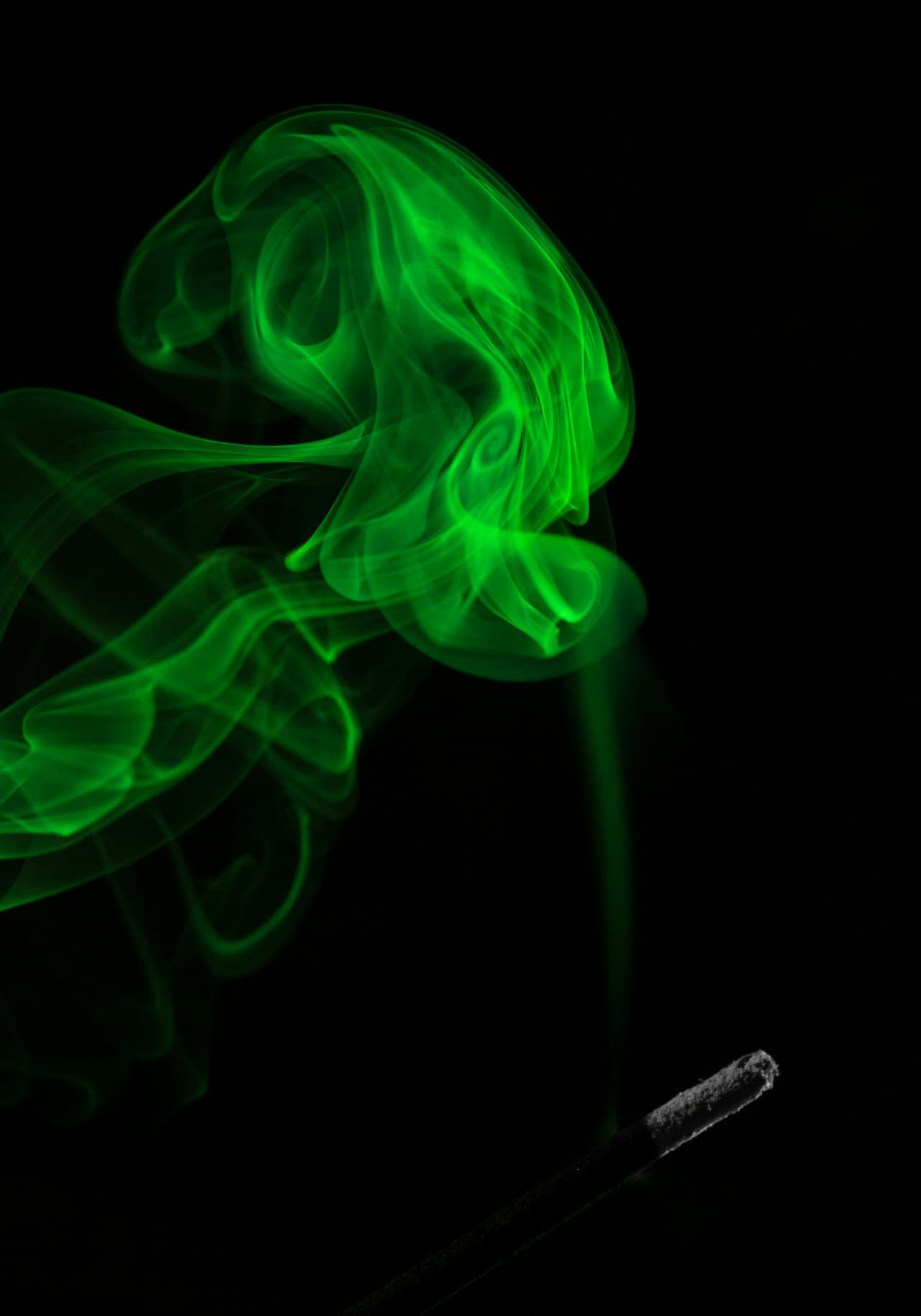 A low-key photograph capturing a stream of neon-green smoke rising from a small smoking stick or cigarette at the bottom edge. The smoke twists into layered curls and translucent sheets, glowing against a pitch-black background. The composition is simple and vertical, focusing attention on the fluid motion and fine texture as the plume expands upward. Lighting is dramatic and directional, making the green appear almost luminous while keeping the surroundings in shadow. The overall mood feels edgy and atmospheric, like a darkroom experiment in color and motion.
