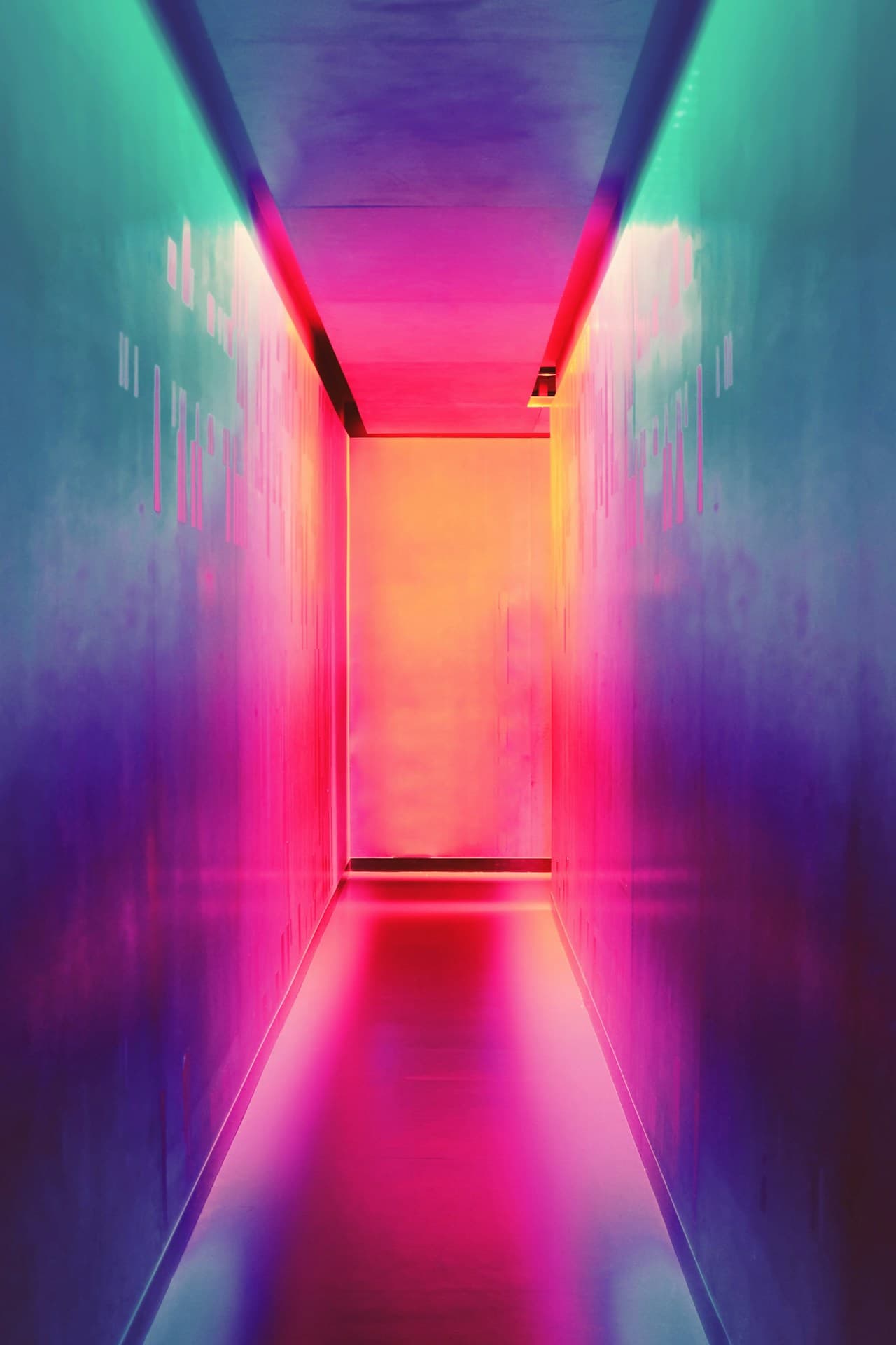 A vivid, futuristic corridor lit with saturated colored light, photographed straight-on in perfect one-point perspective. The narrow hallway has smooth walls that fade from teal and cyan into deep blue and purple, while the ceiling glows violet. At the far end, a warm yellow-orange light source fills the doorway area, creating a strong color contrast with the cool sides. The floor reflects intense magenta and pink tones, forming a bright path toward the vanishing point. The scene feels like a light installation or stylized architectural interior, with a dreamy, synthetic atmosphere and a bold, immersive mood.