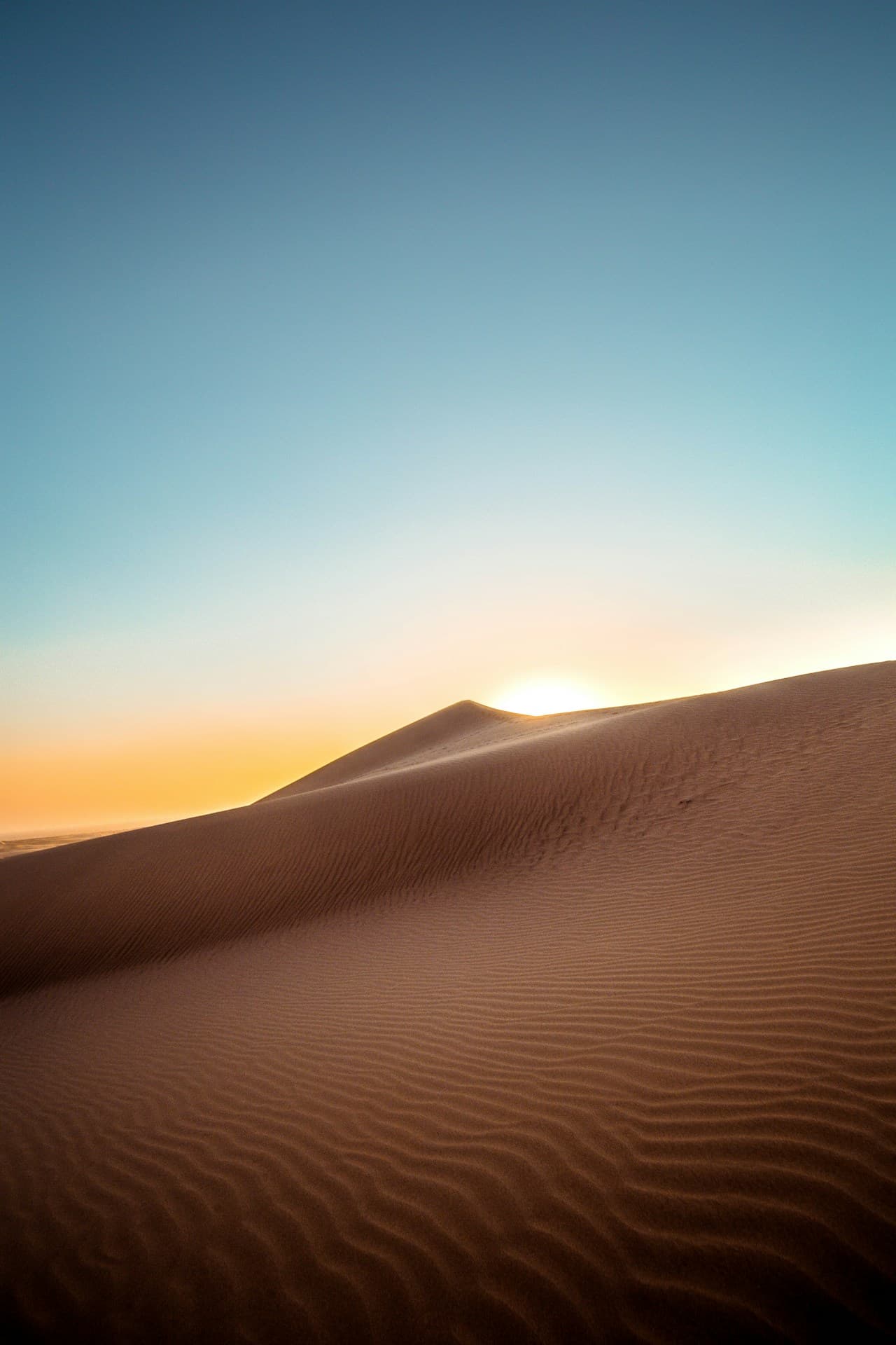 Sunrise over rippled dunes, captured from a low vantage point. The sun sits just behind a distant ridge, casting a warm glow along the horizon while the sky transitions into cool blue above. In the foreground, fine sand ripples create repeating lines and gentle shadows, leading the eye toward the light. The composition balances a large open sky with a sweeping dune slope, keeping the scene uncluttered. Lighting is golden and soft, emphasizing texture without harshness. Mood is peaceful and hopeful.