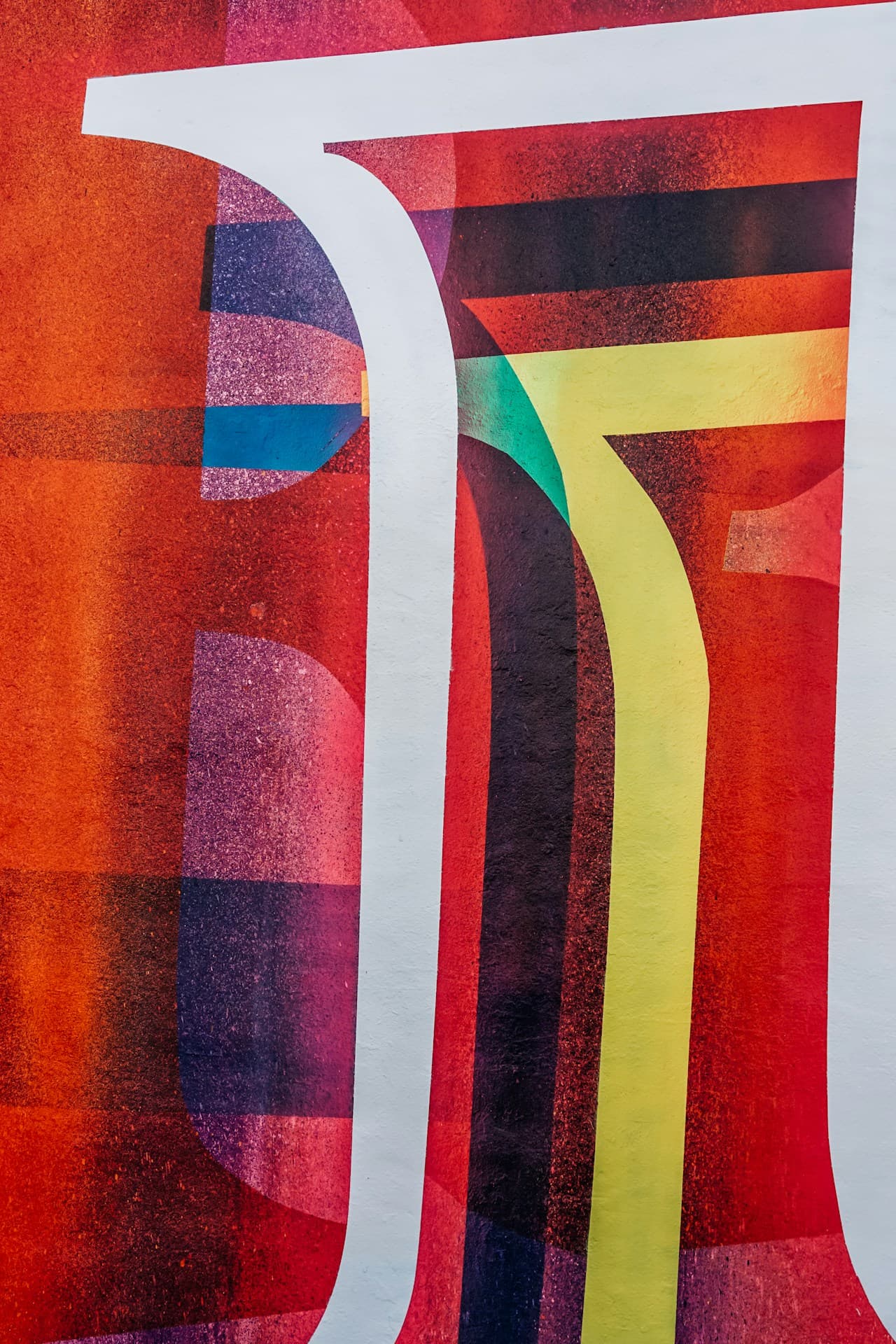 Close-up of a textured painted surface featuring bold graphic shapes. A thick white curved stripe slices through a field of saturated reds and oranges, while layered translucent blocks of purple, teal, and warm yellow overlap in the background. Fine speckling and wall texture are visible, giving the colors a gritty, printed feel. The crop is tight so the shapes feel larger-than-life, with strong vertical and curved lines. Lighting is even and color-forward. Mood is vibrant and urban.